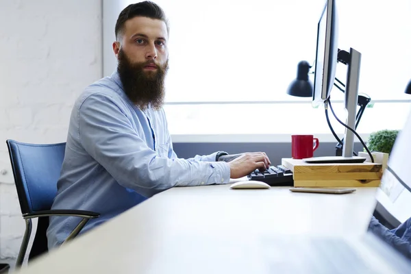 Half length portrait of  bearded employee looking at camera while browsing information in websites for work.Young hipster guy dressed in trendy clothing typing on keyboard sitting in office