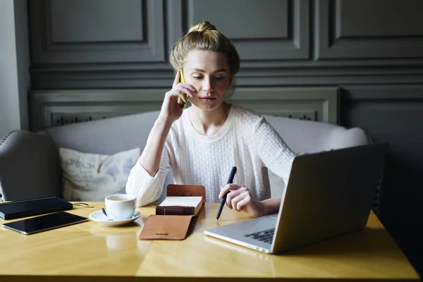 ラップトップ コンピューターを見ながら Wifi に接続された電話にモバイルで会話を有する思いやりのある実業家 居心地の良いカフェに座っている近代的なデバイスの操作魅力的なブロンドの女性 — ストック写真