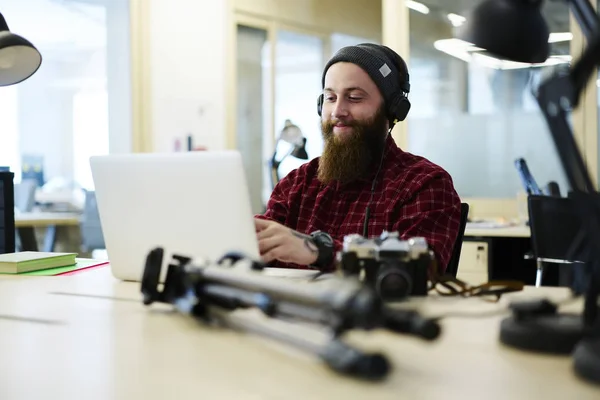 Positive male creating music using application on laptop computer and enjoying made composition via headphones.Bearded man in cool hat listening favorite playlist sitting in coworking