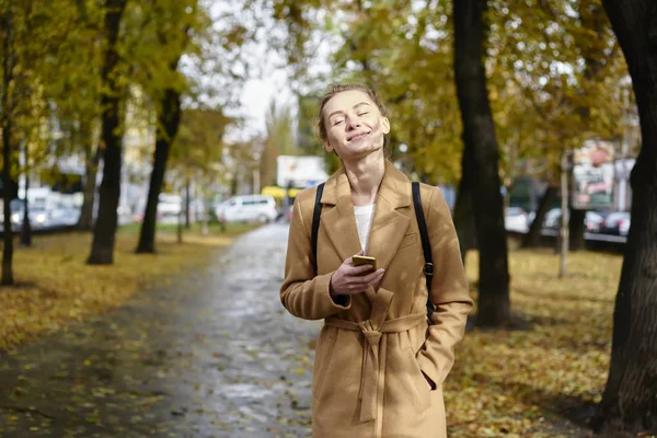 Beautiful female student dressed in stylish brown coat walking in central park and enjoying sunny autumn weather.Attractive hipster girl strolling in urban settings with modern phone in hand