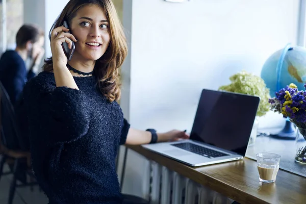 Positive attractive female graphic designer dressed in stylish clothing looking away and communicating with friends using application on cellphone connected to 4G while sitting in coworking