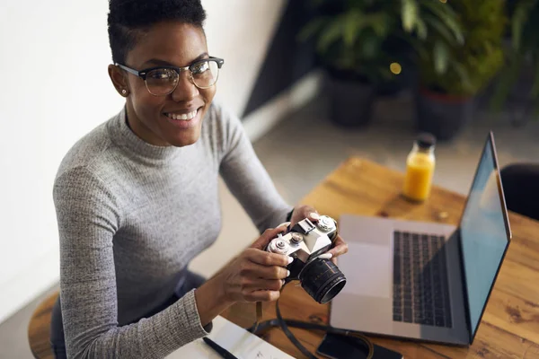 陽気なプロ アフロ アメリカン女性フリーランサー ビンテージ カメラ コワーキング スペースに座っている機器の設定を確認して幸せな写真家によって行われたお金オンライン編集写真を獲得の肖像画 — ストック写真