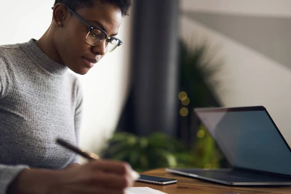Concentrated afro american businesswoman analyzing details of contract before signing sitting in coworking space, pensive web designer making drawings on paper for modeling on laptop computer