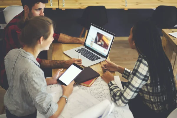 設計図と現代の Macbook コンピューター木製デスクトップに座って新しいグラフィックを作成する議論する若手デザイナー タッチ パッドを使用する作業の多様性グループ 空白の画面領域 広告エリア — ストック写真