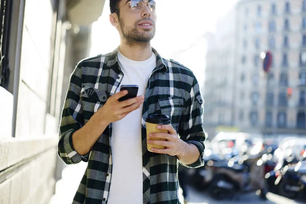 Handsome tourist in cool eye glasses searching direction while strolling in city street with gadget and coffee in hands.Bearded thoughtful man using cellphone maps in urban setting