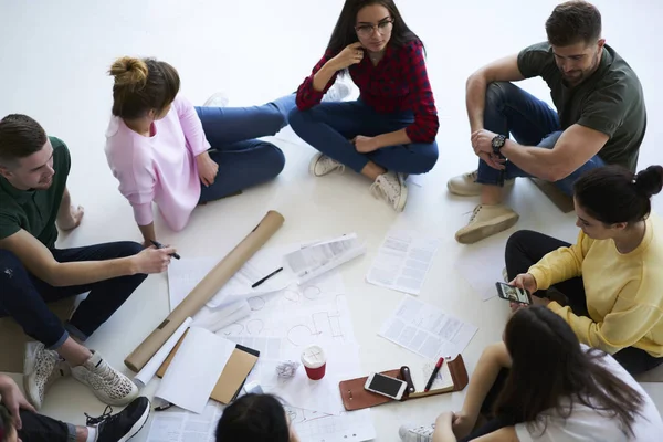 From above view of young coworkers sitting on floor in circle and collaborating on new project.  Creative team of members brainstorming together while discussing project ideas sitting near blueprints