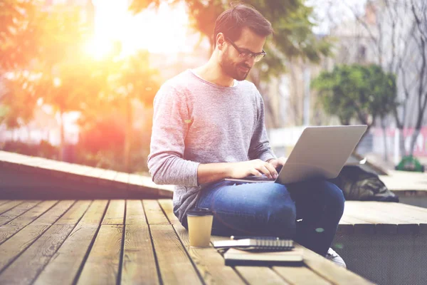 正胡子的时髦的家伙在光学眼镜坐在校园内与咖啡 并键入短信在现代笔记本电脑键盘上使用高速4G 互联网连接 — 图库照片