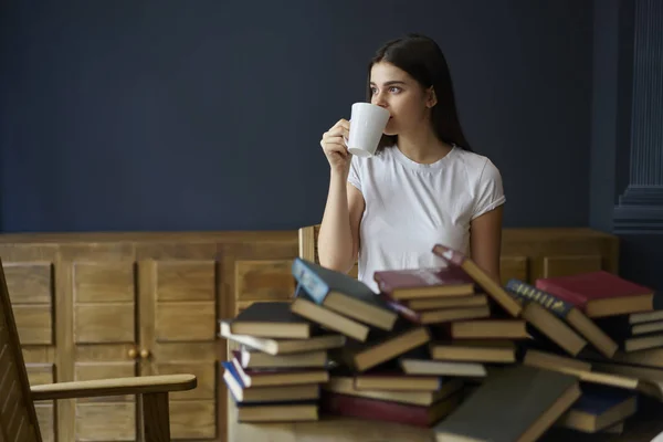 Young attractive student looking away while enjoying aroma coffee sitting in library against wall with promotional background.Pondering beautiful female spending free time with literature indoors