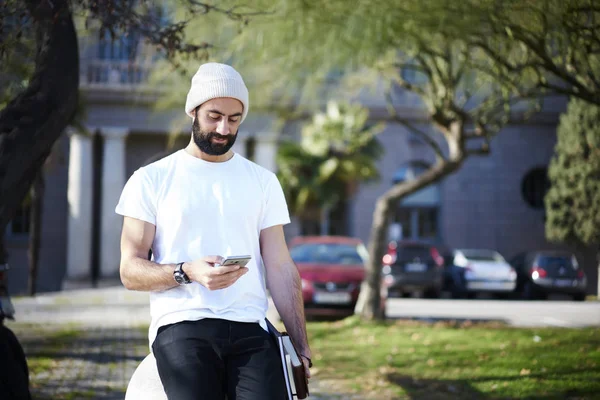 Handsome male traveler strolling on city street using application with online maps on smartphone for getting to destination, hipster guy in casual wear chatting in social networks via 4G connection