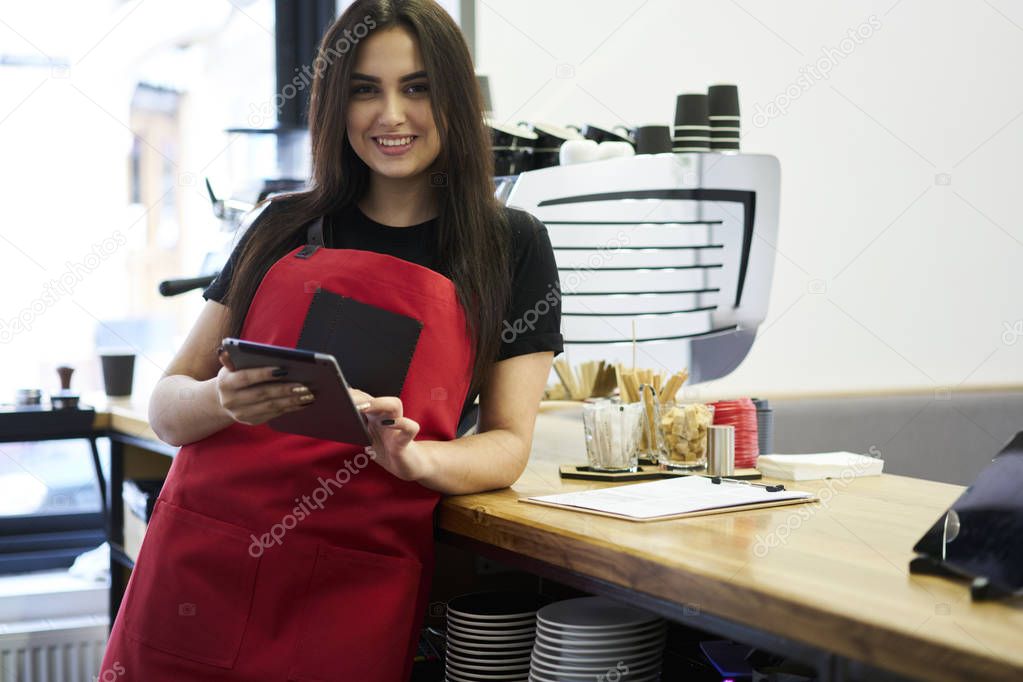 Portrait of attractive young barista checking financial reports on modern touch pad while smiling and looking at camera.Beautiful positive cashier in red apron standing at bar with tablet in hands