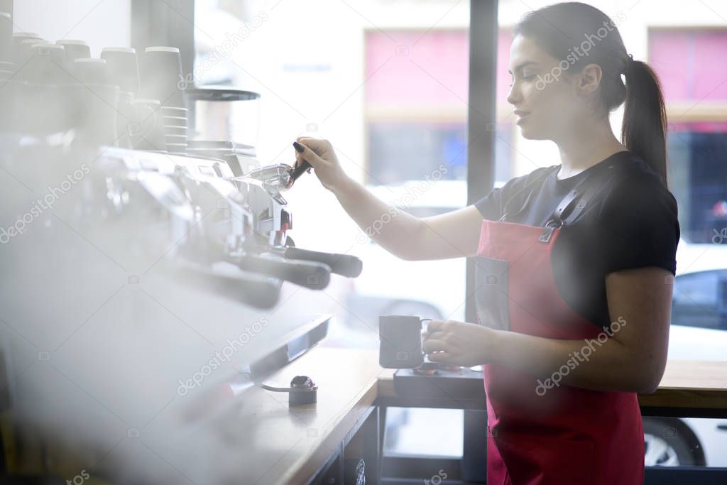 Professional  barista in uniform making aroma coffee using machine concentrated on working process.Beautiful brunette waitress preparing cappuccino doing order of cafeteria client standing at bar
