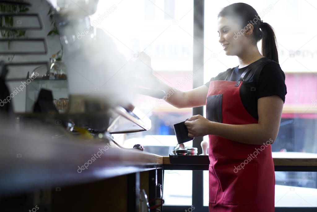 Attractive brunette female barista enjoying working process preparing coffee drinks using professional equipment in cafeteria.Skilled waitress in apron steaming milk on machine making cappuccino