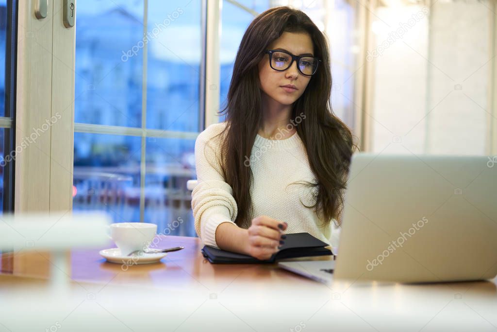 Pensive young brunette employee making researches in network watching videos analyzing information, professional project manager in eyewear working on laptop application having coffee break in cafe