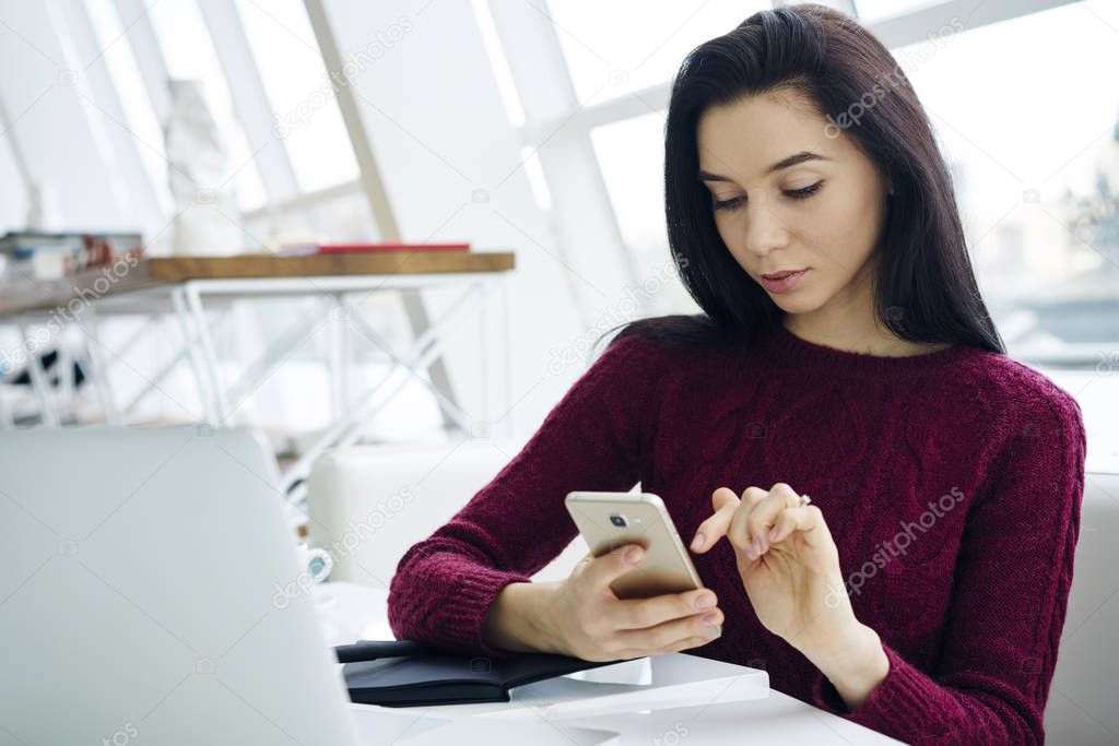 Attractive female installing mobile application on modern smartphone while sitting in stylish office.Hipster girl reading received sms message on cellular.Copy space area for your advertising text