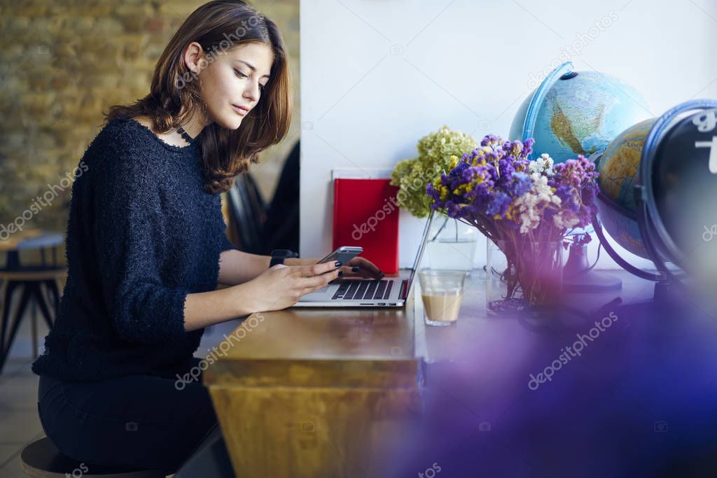 Charming young female student watching interesting video on smartphone connected to 4G internet while preparing for coursework project.Concentrated young woman texting feedback for website indoors