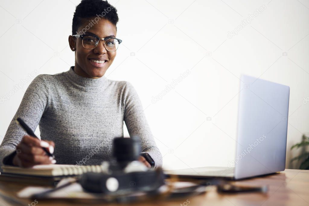 Portrait of attractive cheerful afro american buinesswoman in glasses satisfied with occupation making distance job in coworking office planning financial report sitting on white promotional background