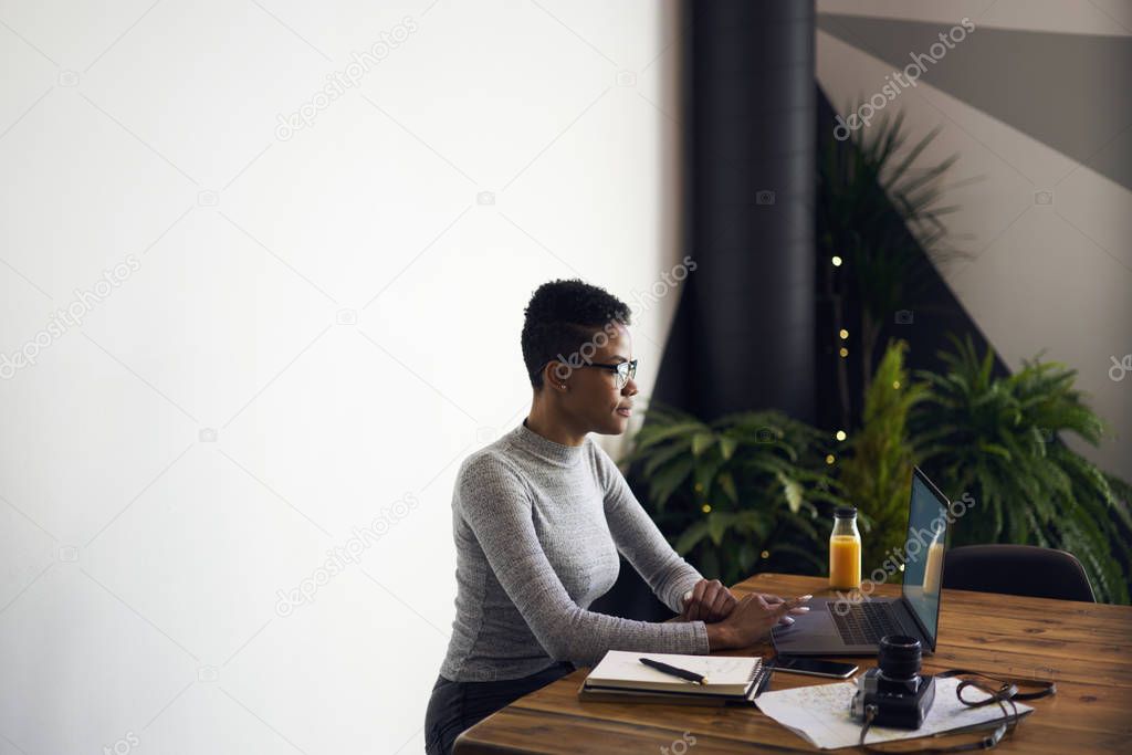 Prosperous afroamerican female owner of trading company  browsing information for developing marketing strategy sitting on white promotional background, skilled freelancer downloading files on laptop