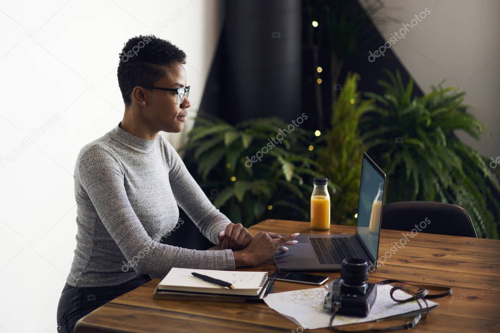 Prosperous afroamerican female owner of trading company  browsing information for developing marketing strategy sitting on white promotional background, skilled freelancer downloading files on laptop