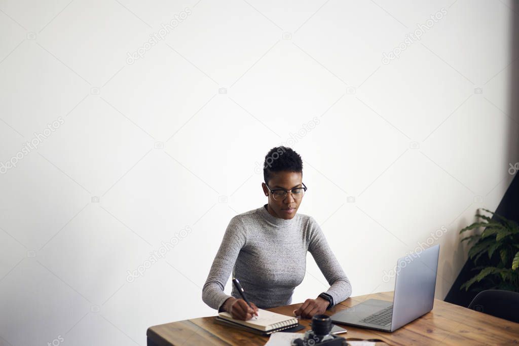 Concentrated Afro-American female designer creating sketch in notebook concentrated on ideas, professional businesswoman in eyewear planning projects making notes sitting on promotional background