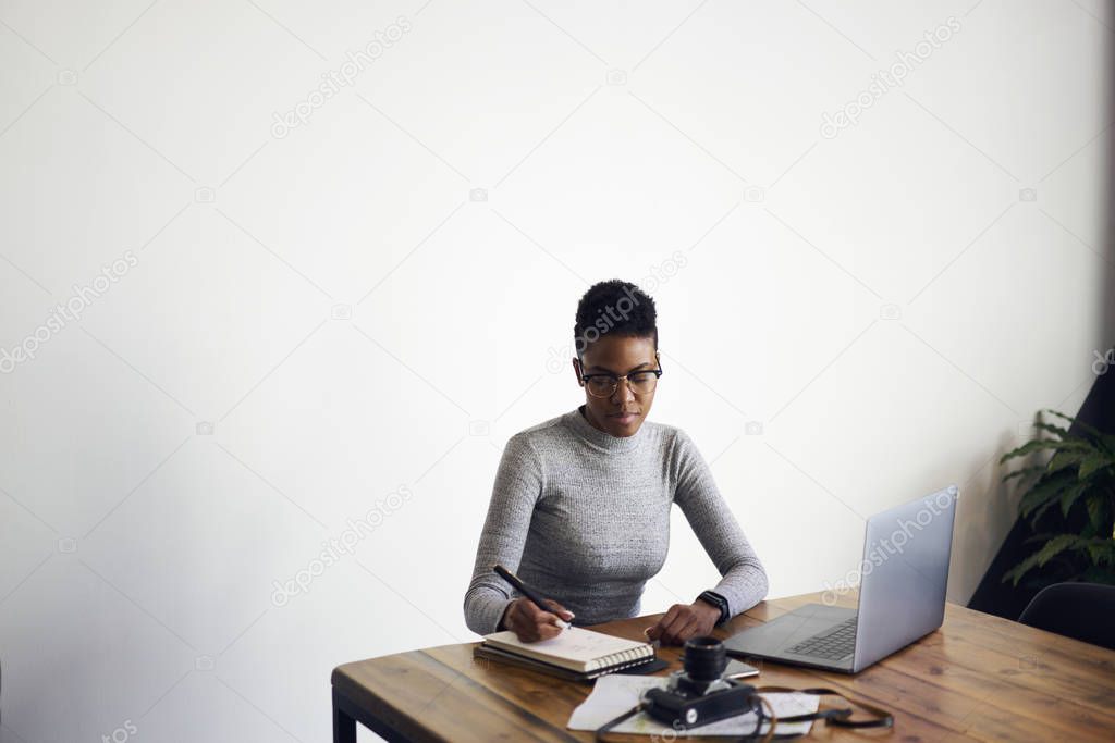 Concentrated afro american businesswoman writing planning of working process in notebook, skilled designer in eyewear drawing sketch for project sitting on copy space area for your advertising 