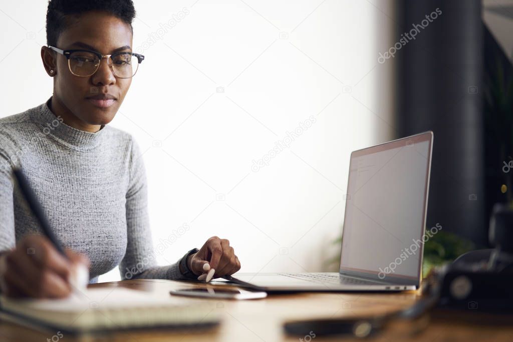 Prosperous afro american businesswoman concentrated on developing strategy for project making accountings and analyzing income making notes while sitting near laptop computer with mock up screen 