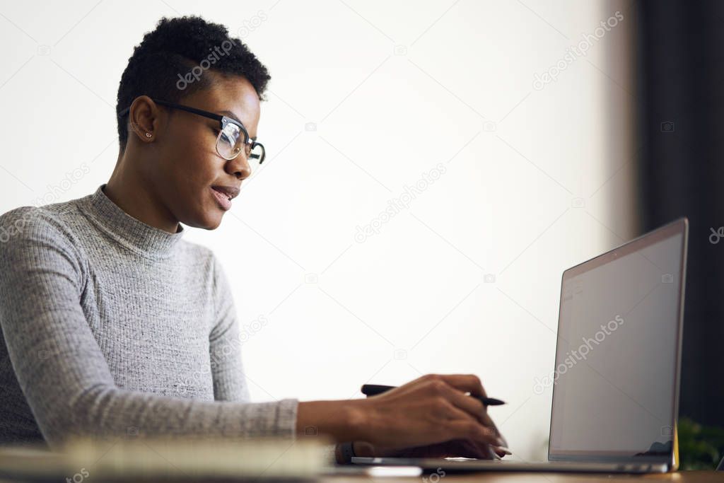 Concentrated afro american freelancer in eyewear earning money online completing task with updating content on websites using laptop computer with mock up screen sitting near copy space for advertising