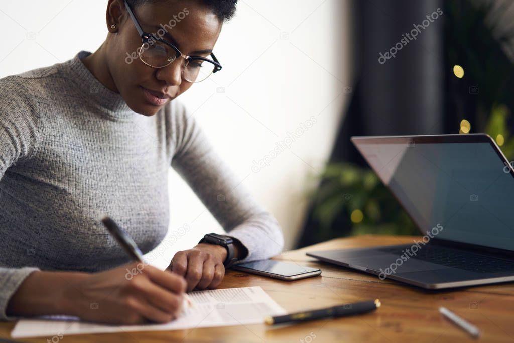 Prosperous afro american businesswoman writing report about company income analyzing information sitting near laptop computer, serious freelancer in eyewear planning working process and tasks