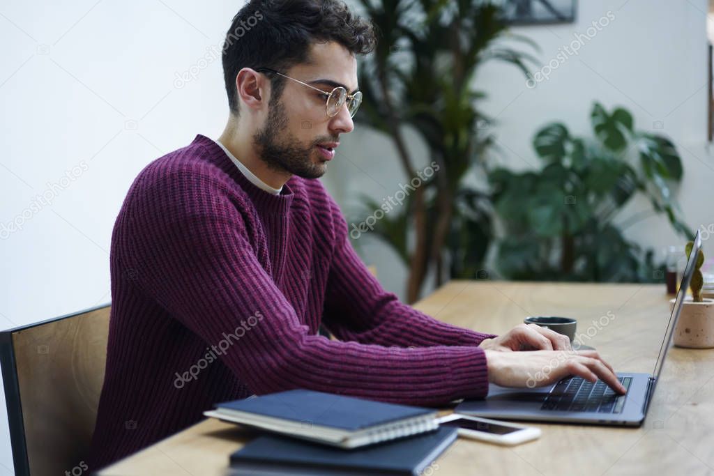 Pensive young male designer in eyewear working on creating graphic on modern laptop device connecting to wireless internet.Concentrated IT developer keyboarding text on computer sitting in office