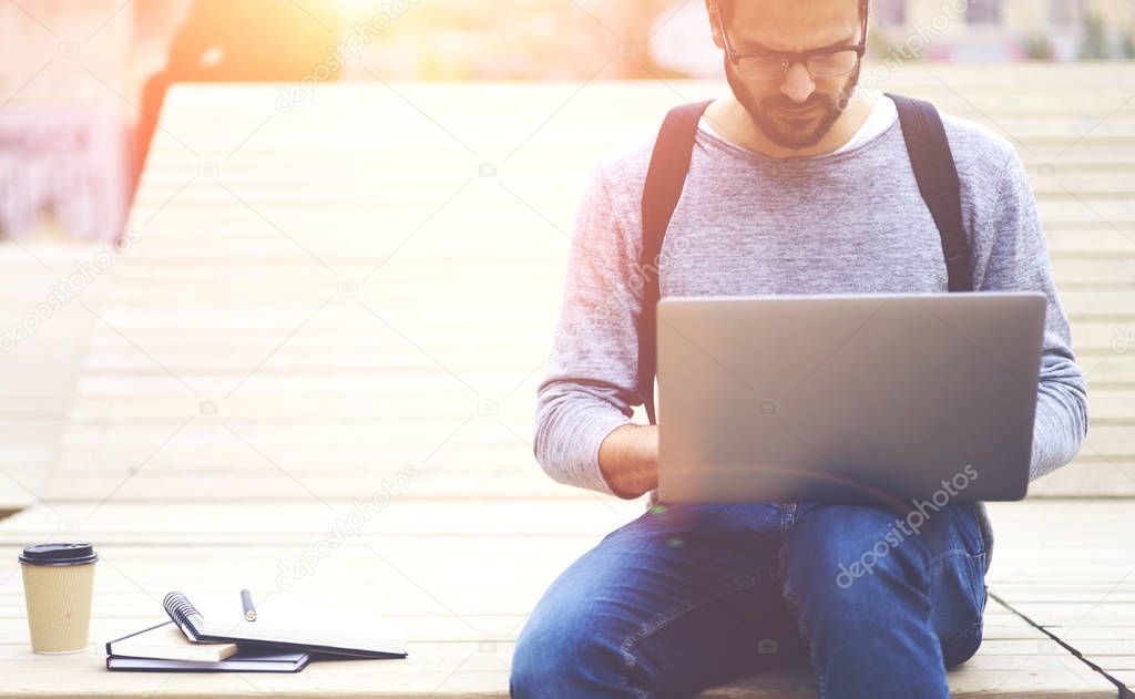 Concentrated male tourist in eyeglasses searching information on laptop computer connecting to wireless 4G internet connection sitting in urban settings.Copy space area for advertising content