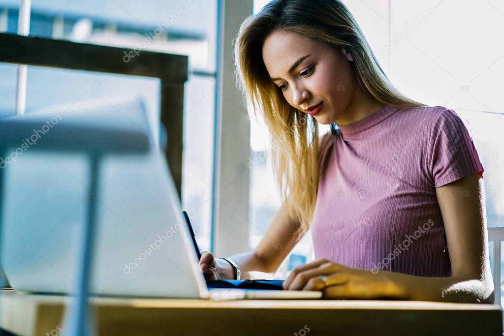 Concentrated blonde female student writing report doing homework using technology for searching information, attractive clever journalist pondering while creating article sitting in coworking office