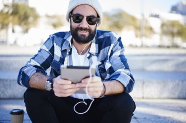 Neşeli sakallı adam trendy güneş gözlüğü ve eğlence açık havada kentsel ayarları süre dinleme şarkıları kulaklık yayın radyo dijital tablet elektronik kitap okuma zevk kıyafeti