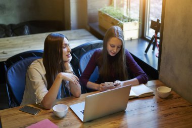 İki hippi kız birlikte şık kafede macbook bilgisayar kitapları ile oturma alanı coworking eğitim. İletişim ve sınava hazırlık sırasında cep telefonu mesajlaşma gençler