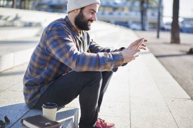 Yakışıklı adam rahat kıyafet bloglama smartphone ve 4 g internet bağlantısı kullanma sosyal ağlarda gülümseyen, güneşli gün boyunca şehir meydanında otururken arkadaşlarınızla sohbet hipster adam sakallı
