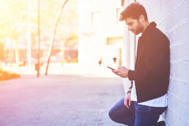 Şık giyim açık havada duvara yakın serbest zaman harcama ve takipçileri smartphone uygulama kullanarak sosyal ağlar ile sohbet ederken duran yakışıklı sakallı öğrenci giyinmiş