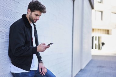 Kentsel ayarda gezinme sırasında bildirim modern telefon üzerinde kontrol iyi görünümlü kendine güvenen bir adam. Genç hipster adam teknoloji ayakta beyaz duvara kullanarak arkadaşlarınızla iletişim