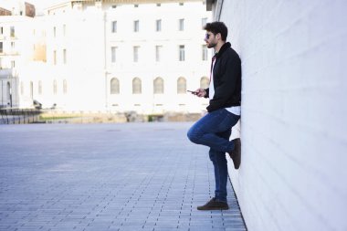 Yakışıklı sakallı blogger giymiş şık kıyafet manifatura ve sosyal ağlar üzerinde modern telefon iletileri gönderme bağlı 4 g için Internet güneşli kentsel ayarı duvara yakın dururken