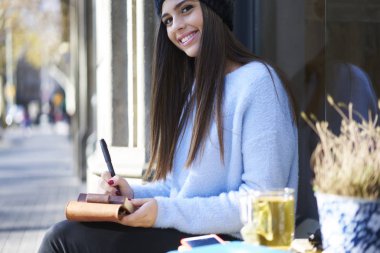 Kameraya bakarak ve bazı notlar not defteri açık havada kahve dükkanında otururken yaparken gülümseyen güzel gazeteci portresi kırpılmış. Çekici muhabiri Not defteri bilgileri yazma
