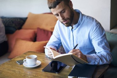 Yakışıklı erkek gazeteci seyredebileceğiniz çalışma planı alanı iç coworking içinde kahve masada oturan defter. Genç yetenekli yazılı Kayıt Defteri'nde yeni romanı için mola sırasında kapalı yazar.
