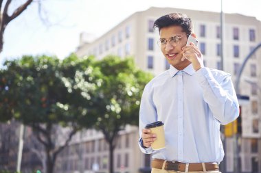 Gülümseyen yakışıklı genç kentsel ayarda yürüyüş sırasında Telefonda komik konuşma yapıyor. Pozitif genç hipster adam akıllı telefon aracılığıyla arkadaşlarınızla konuşurken ücretsiz anda şık gözlükler