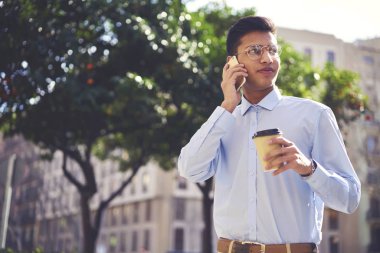 Genç olumlu hipster adam cep telefonu üzerinde konuşurken kablosuz internete bağlıyken şık kıyafet uzağa bakarak giyinmiş. Yakışıklı genç lezzetli içecek açık havada serbest zaman zevk