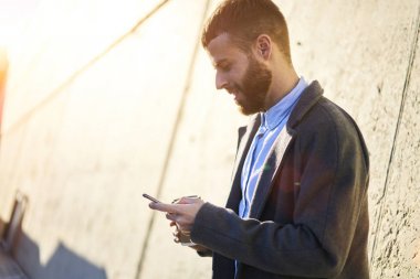 İyi bir ruh hali ile 4 g bağlantı ayakta kahve ile kentsel ortamda modern cep üzerinde arkadaşlarınızla çevrimiçi sohbet ile genç gülümseyen kişi. Metin iletiniz için yer alan kopyalamak veya reklam