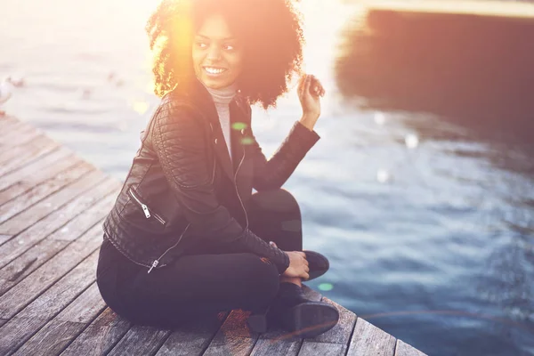 Pondering happy gorgeous woman dressed in trendy clothing looking away during resting on wooden pier near to lake at sunset time.Smiling young attractive hipster girl having recreation outdoors