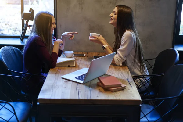 Two best friends talking about funny stories and enjoying leisure time with tasty coffee in stylysh cafeteria.Teenagers sitting in coworking with laptop device and communicating with each other