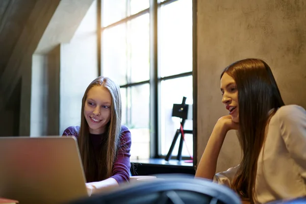 Cheerful young women watching funny videos in social networks on modern laptop device connecting to free internet.Smiling hipster girls browsing websites on computer enjoying free time in coffee shop