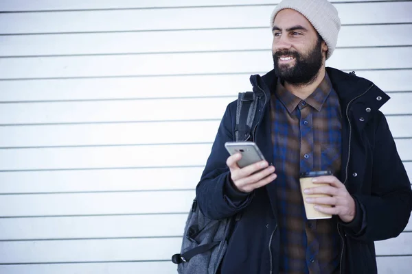 カジュアルな服装とタクシー サービスをプロモーション背景にコーヒーのテイクアウトの インターネット接続に立ってを介してオンライン支払うことを呼び出すアプリケーションを使用して帽子を着て肯定的な流行に敏感な男 — ストック写真
