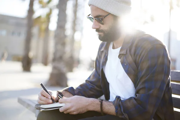 Handsome concentrated bearded male tourist in casual outfit resting on bench near copy space area for your advertising create text of article for blog writing in notepad plans and ideas of trip