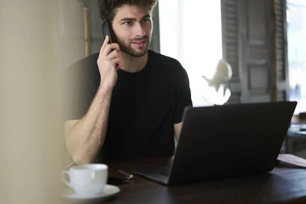 室内のコーヒー ショップでノート パソコンに座って近代的な携帯で同僚と話して夢のような起業家のイメージをトリミングします 距離の仕事中に電話を介して通信男性コピー ライター — ストック写真