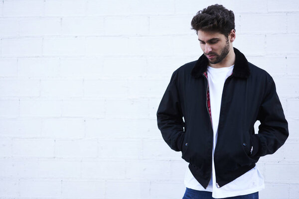Male bearded student dressed in trendy clothing standing near white wall enjoying free time in urban settings.Promotional background for advertising.Copy space area for text message