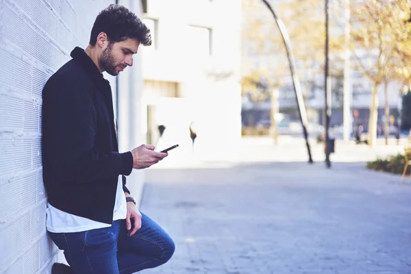 Handsome bearded student dressed in stylish clothing standing outdoors near to brick wall while spending free time and chatting with followers in social networks using application on smartphone