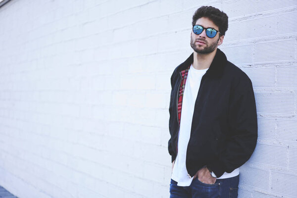 Half length portrait of confident handsome man in stylish sunglasses looking at camera while recreating at urban setting.Bearded young man standing against wall with copy space for advertising text